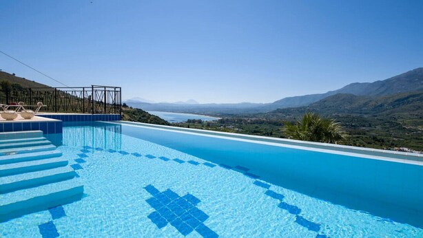 A clear blue swimming pool with steps leading into it, surrounded by a mountainous landscape and a view of a distant body of water.