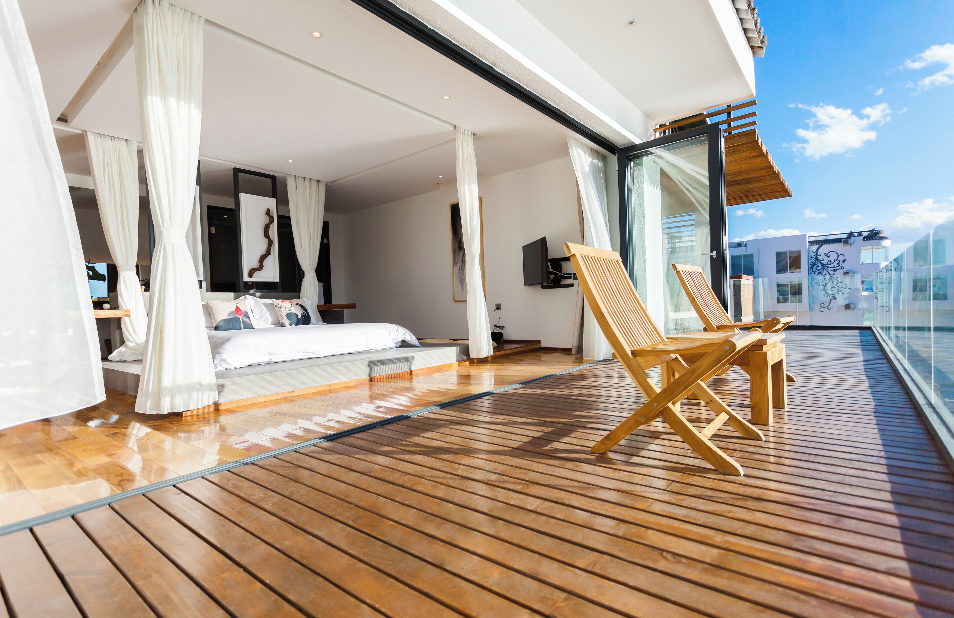 A spacious balcony with wooden flooring, a bed with white bedding, and a seating area with wooden chairs.