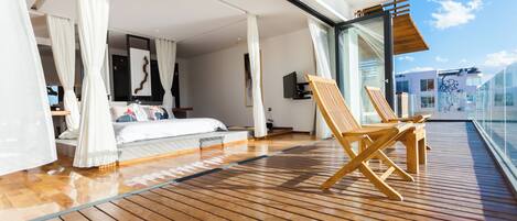 A spacious balcony with wooden flooring, a bed with white bedding, and a seating area with wooden chairs.