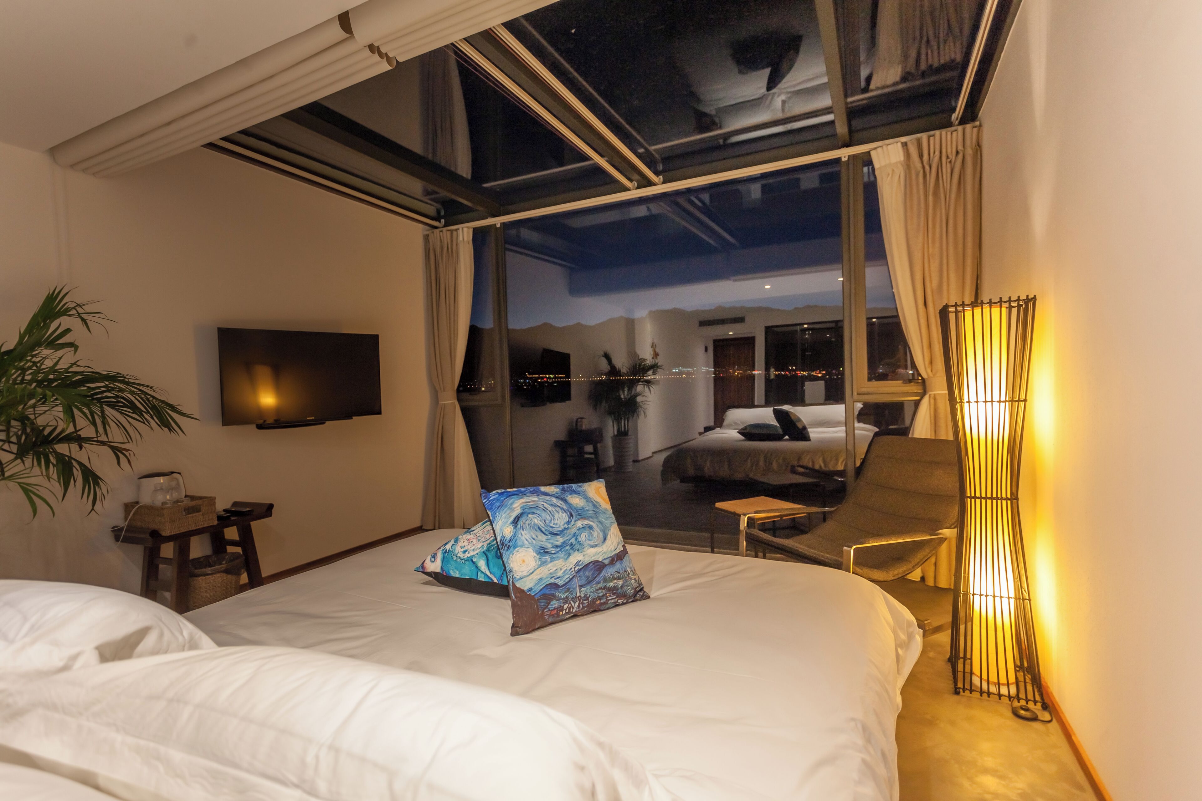 A hotel room with a bed, a chair, a lamp, and a large window offering a view of the city.