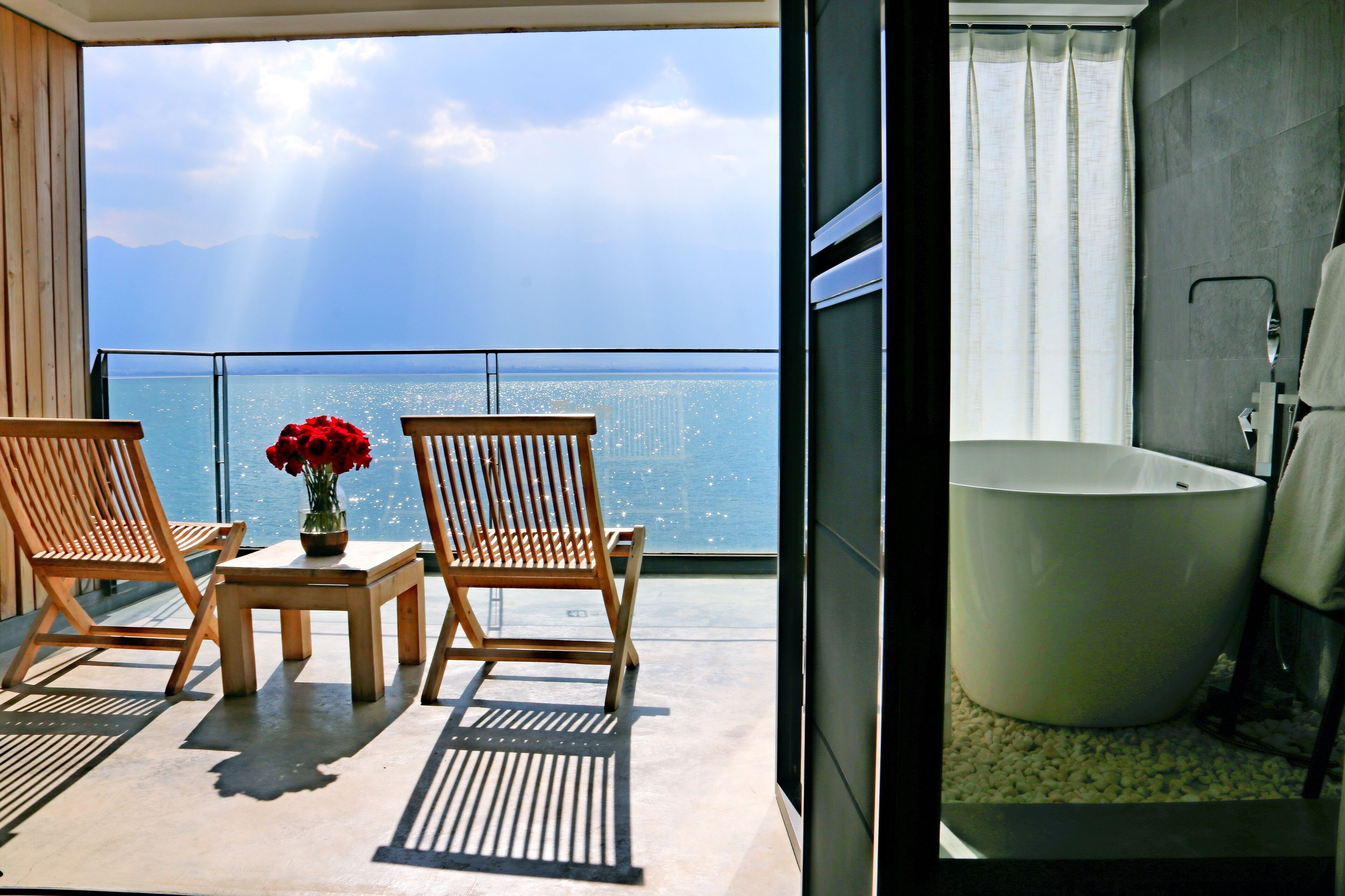 A balcony with two wooden chairs, a small table, and a vase of red flowers.
