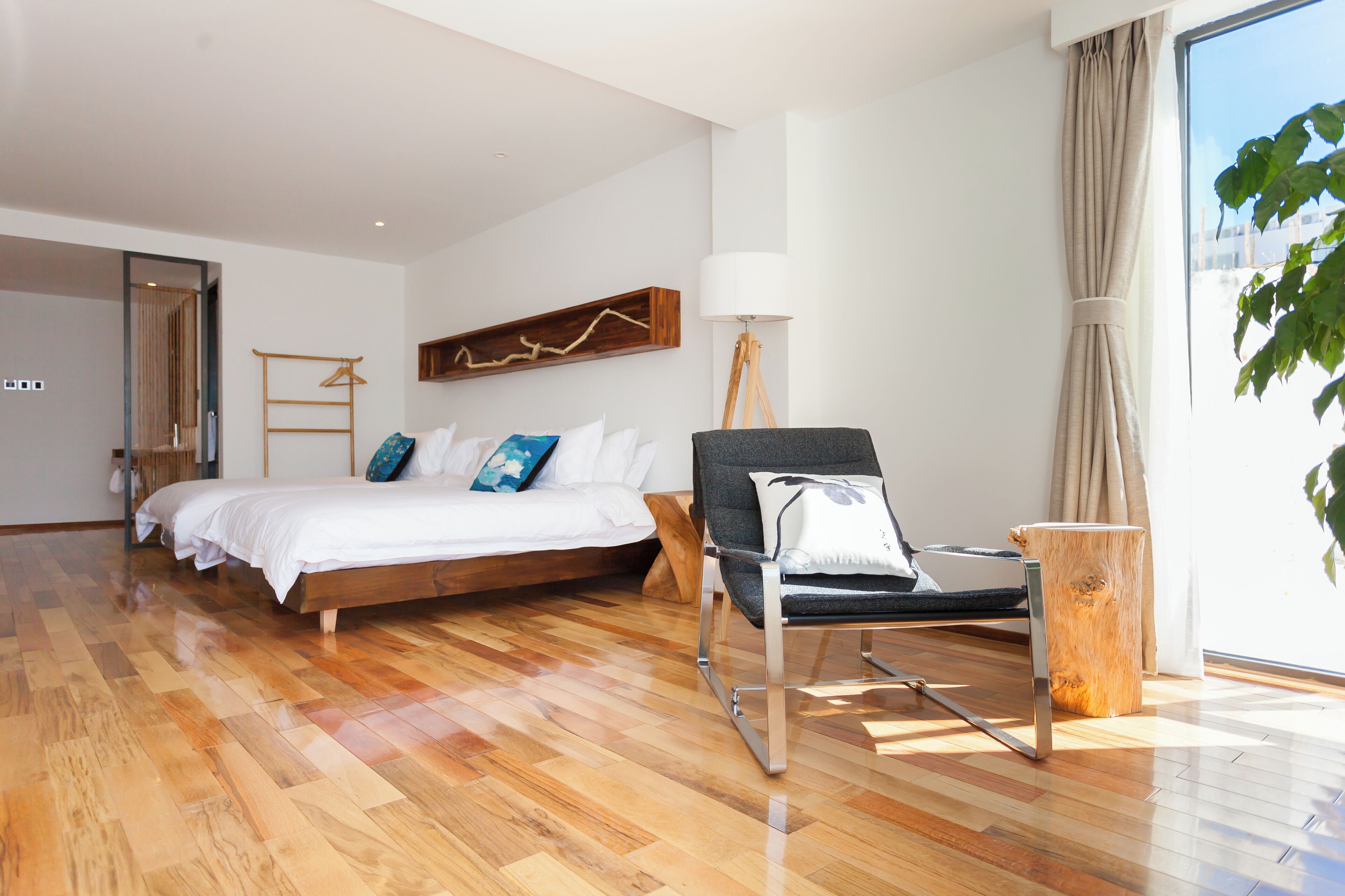 Un dormitorio moderno con piso de madera, una cama con ropa blanca, una silla gris, una mesita de madera y un ventanal con cortinas.