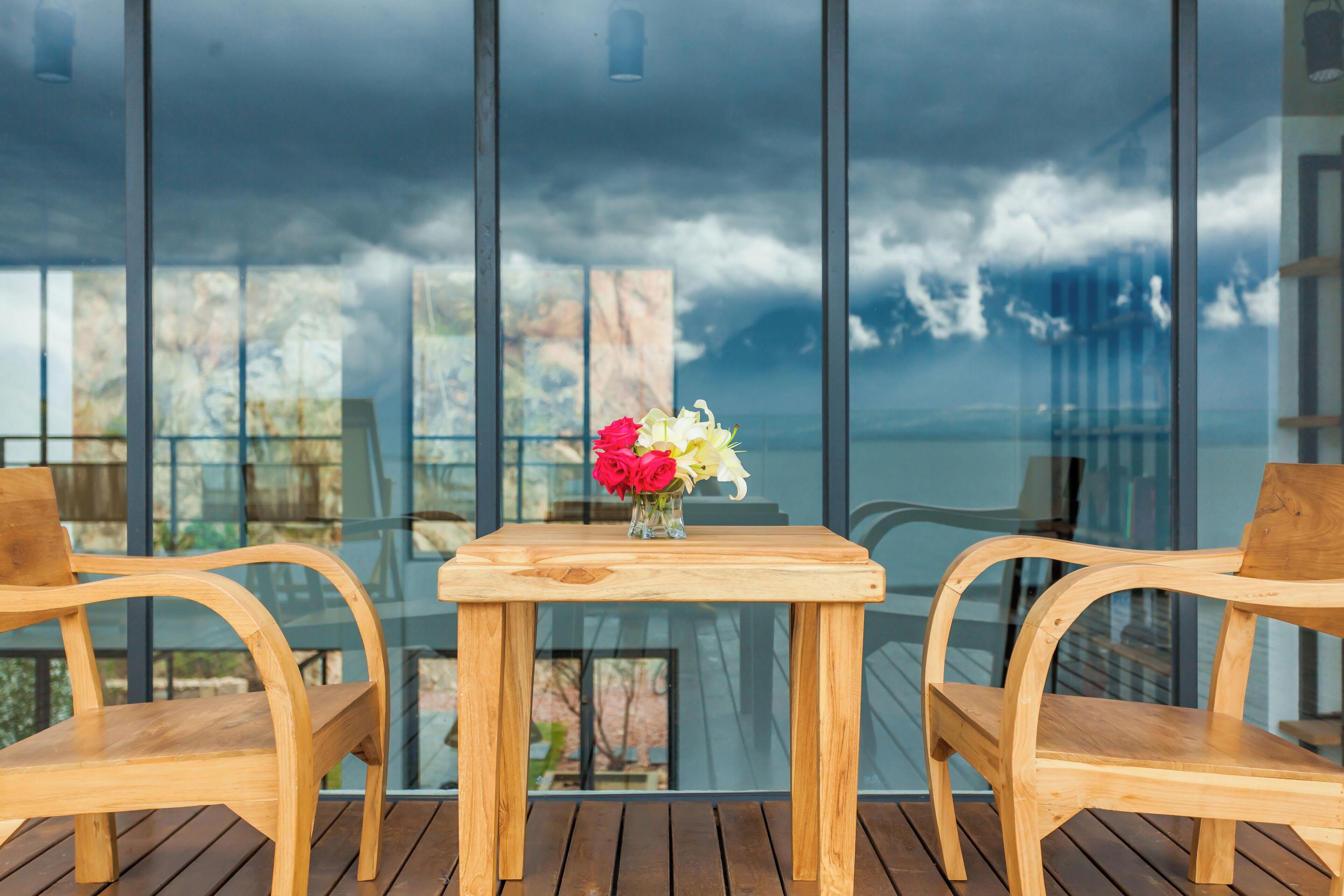 A wooden table with a vase of flowers and two wooden chairs on a balcony with a glass railing, overlooking a scenic view.