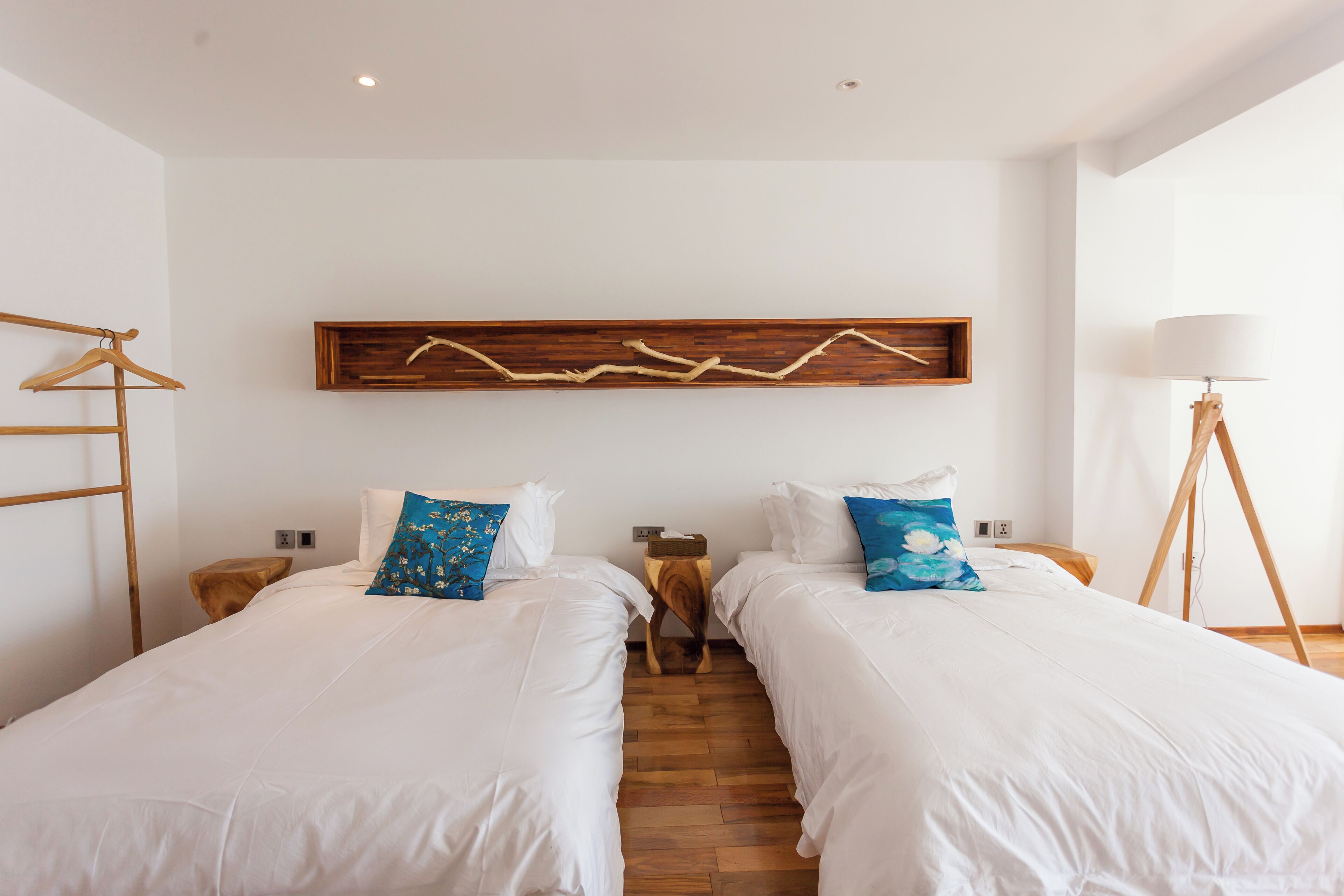 A hotel room with two single beds, wooden headboards, a wooden artwork above the beds, a wooden floor, and a bedside table with a lamp.