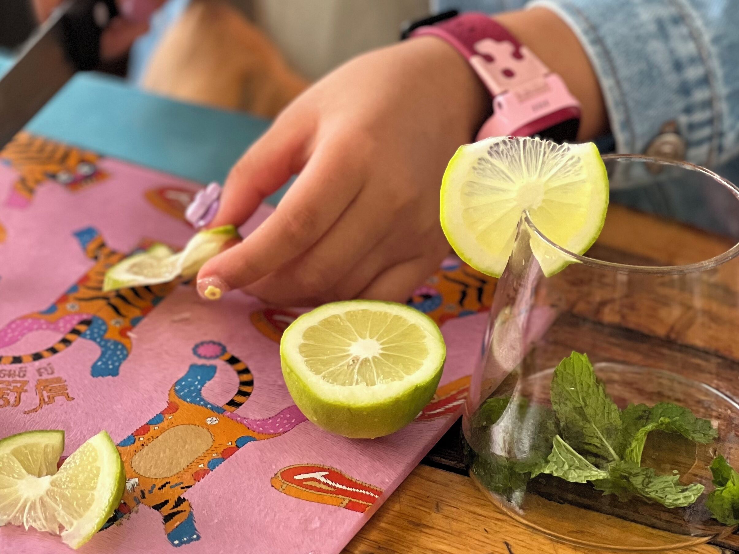 Un tavolo con un sottotavolo colorato, un bicchiere con fette di lime e foglie di menta, e una mano che raccoglie una fetta di lime.