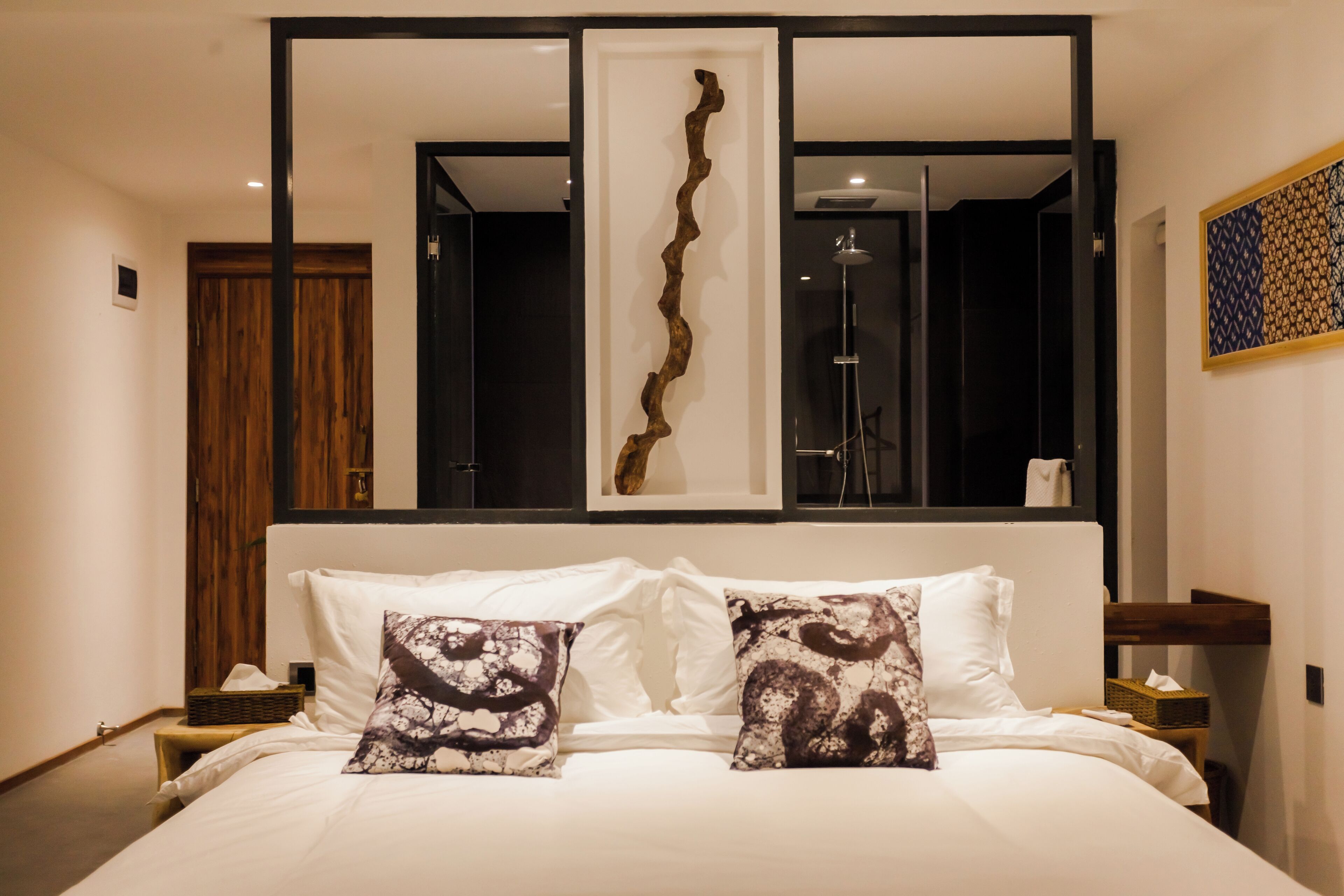 A modern bedroom with a large bed, two decorative pillows, a wooden headboard, a wall-mounted artwork, and a nightstand with a lamp.