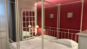 Ein Raum mit roten Wänden, ausgestattet mit einem Himmelbett, weißer Bettwäsche, einem Spiegel und zwei Wandleuchten.
