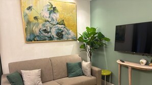 Ein Wohnzimmer mit einem beigen Sofa, grünen Kissen, einem großen Gemälde, einer Zimmerpflanze, einem Fernseher und einem kleinen Tisch mit einer Vase und einer Fernbedienung.