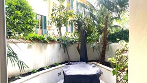 Ein überdachter Außenbereich mit Tisch und Stühlen, umgeben von Pflanzen und einem Gebäude im Hintergrund.