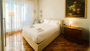 A neatly made bed with white linens and towels, a wooden floor, a chair, a bedside table with a lamp, and a painting on the wall. - Verrazzano House Florence (Firenze)