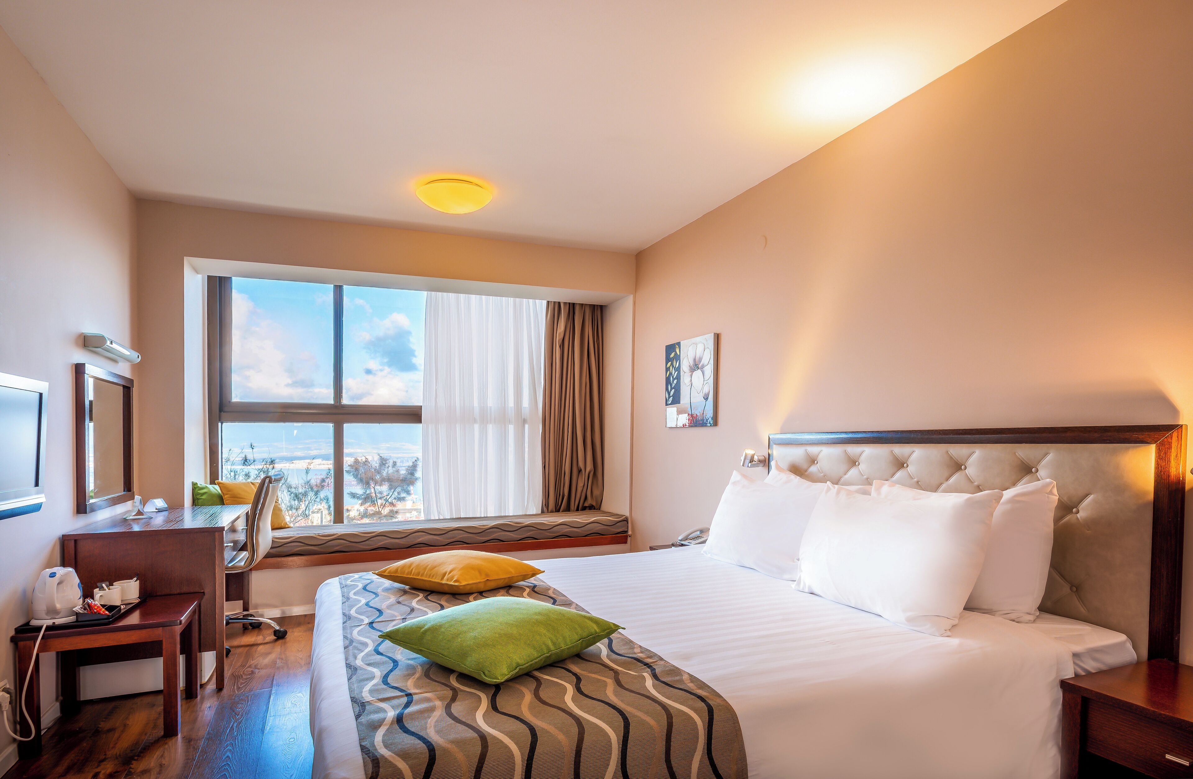 A hotel room with a large bed, a desk with a chair, a window with a view, and a bedside table.