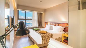 A hotel room with a bed, bedside table, lamp, and a view of the city through large windows.