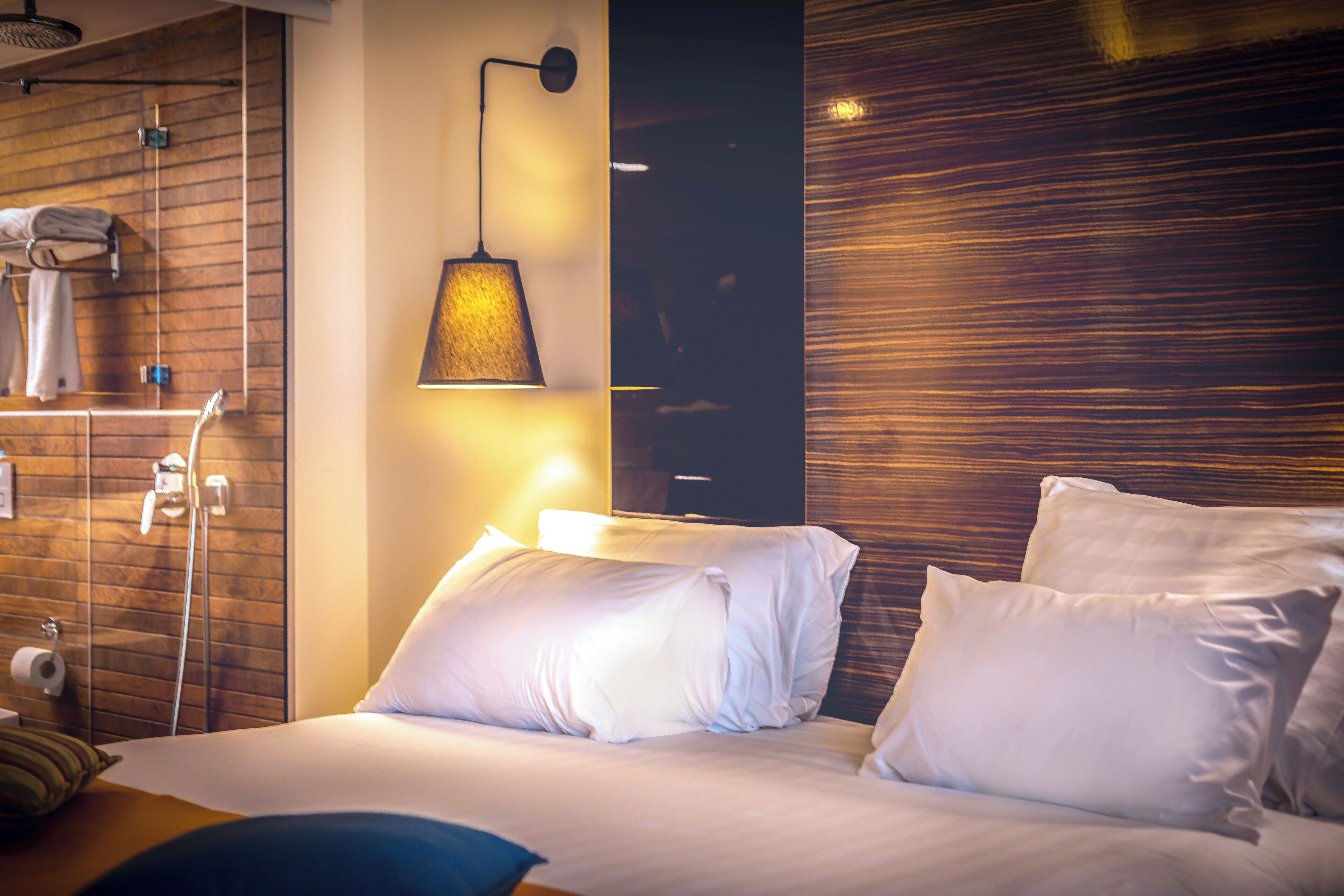 A modern hotel room with a wooden wall, a hanging lamp, a bed with white linens, and a towel rack with towels.