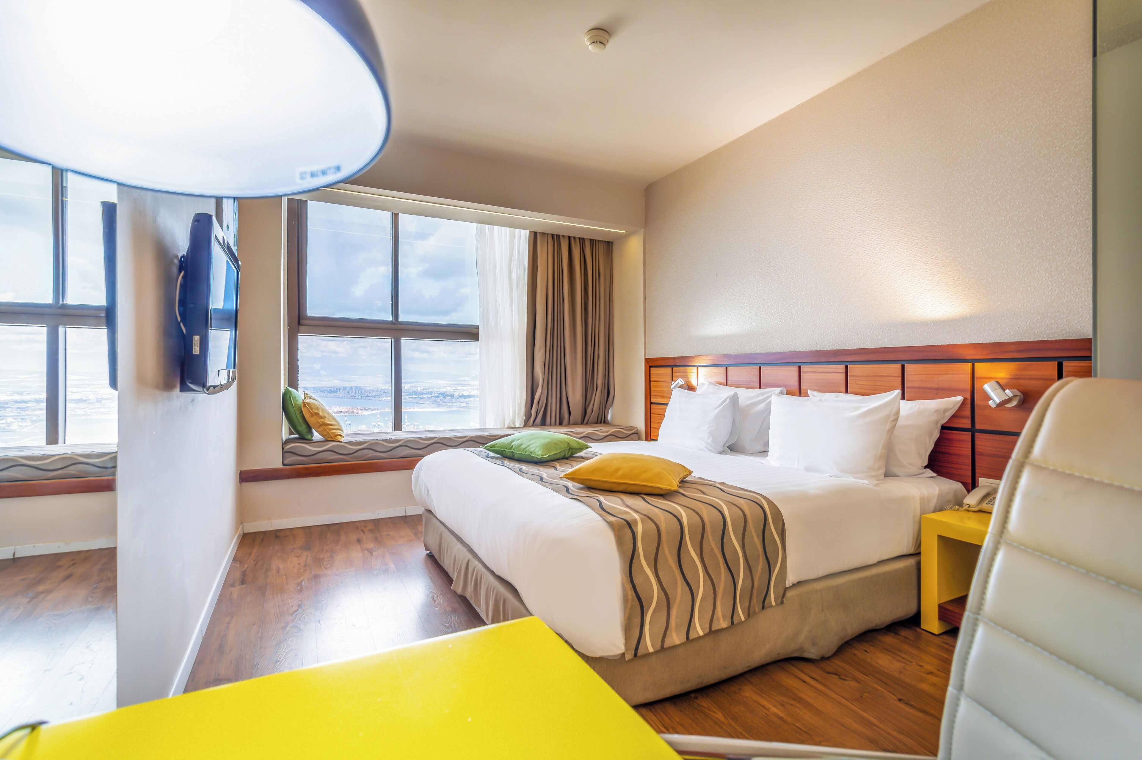 Habitación de hotel con un ventanal grande, una cama con manta a rayas, una mesita de noche con lámpara y una silla.