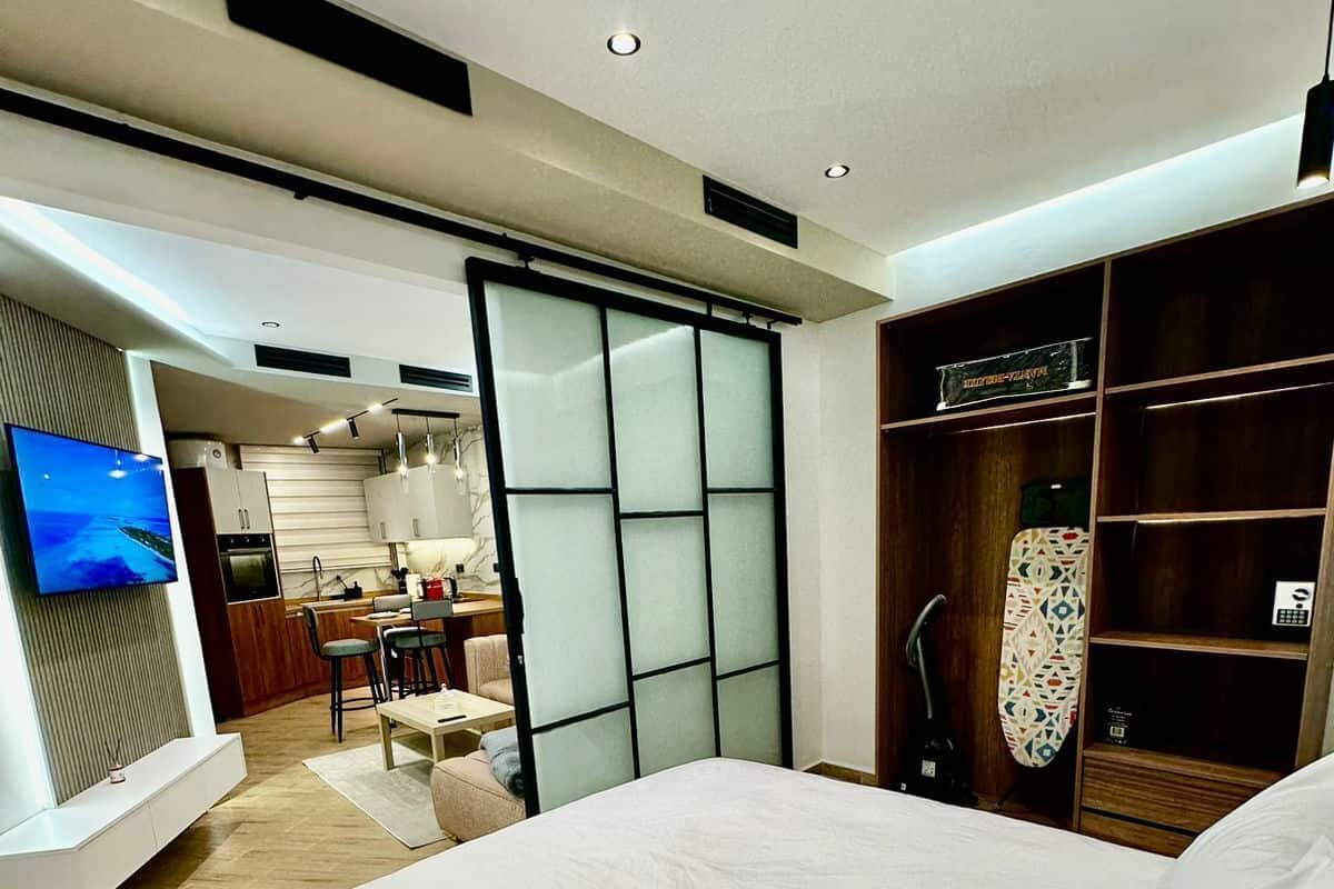 A modern hotel room with a large bed, a TV mounted on the wall, a kitchenette, and a sliding glass door leading to a balcony.