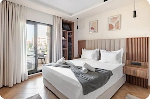 A hotel room with a large bed, a wardrobe, a seating area with a chair, and a view through a glass door.