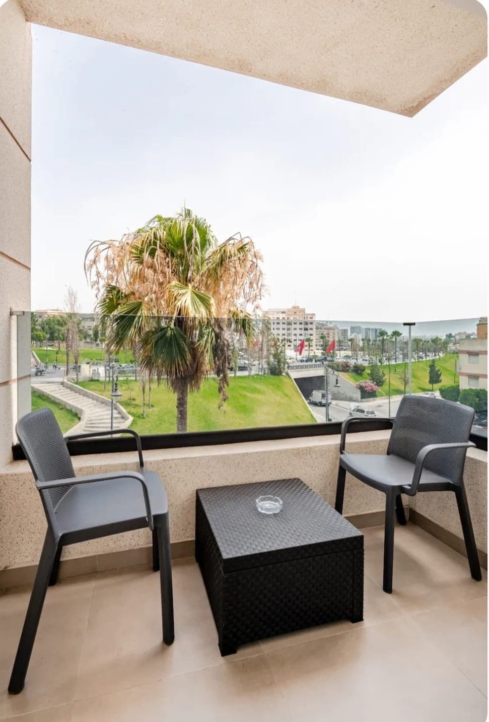 A balcony with two chairs and a small table, overlooking a cityscape with palm trees and buildings.