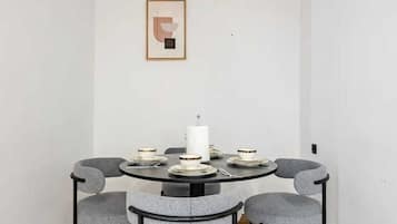 A dining area with a round table, four grey chairs, a white wall with a framed picture, and a hanging light fixture.