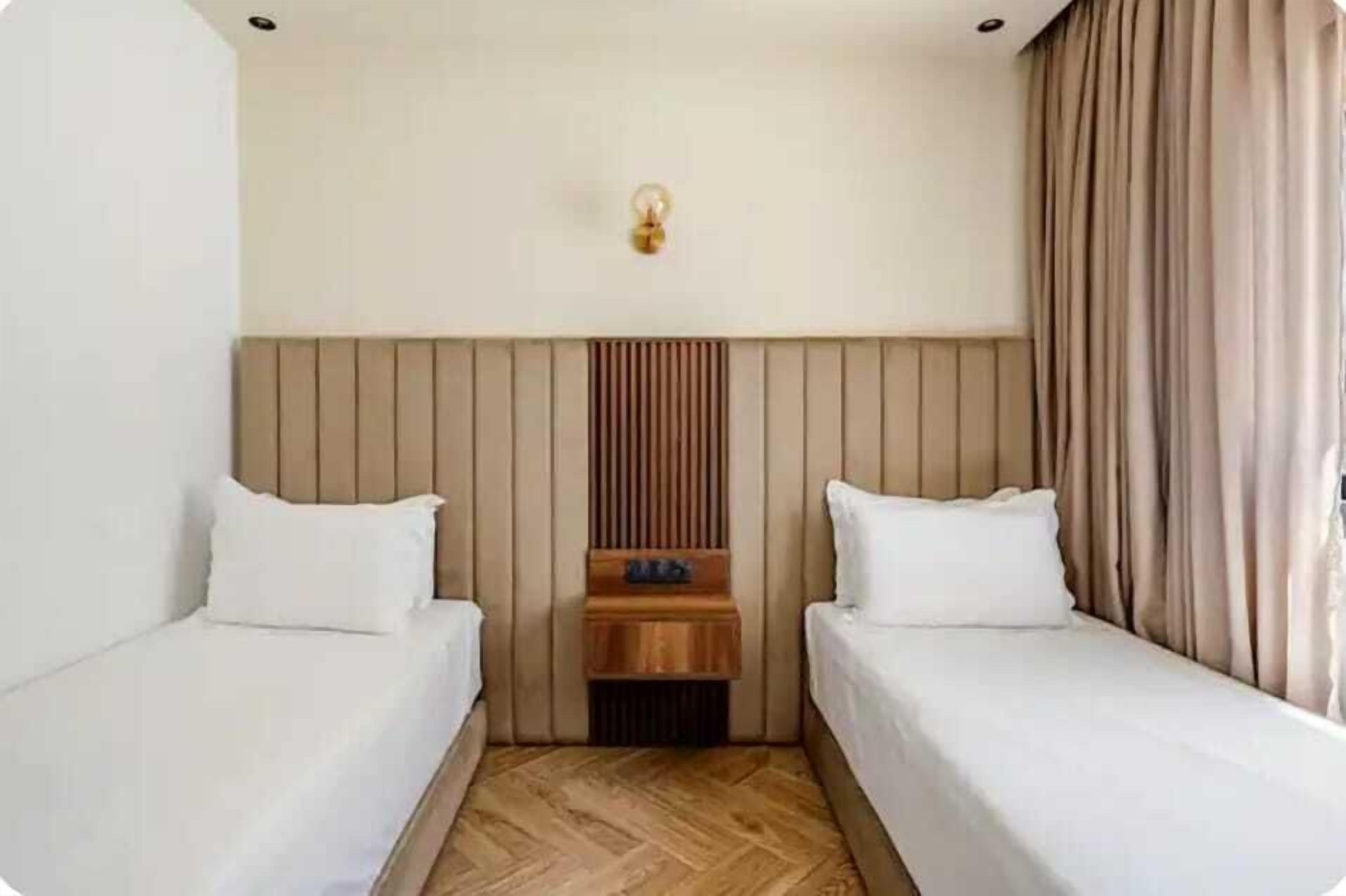 A hotel room with two single beds, a wooden headboard, a nightstand, and a wall-mounted light fixture.