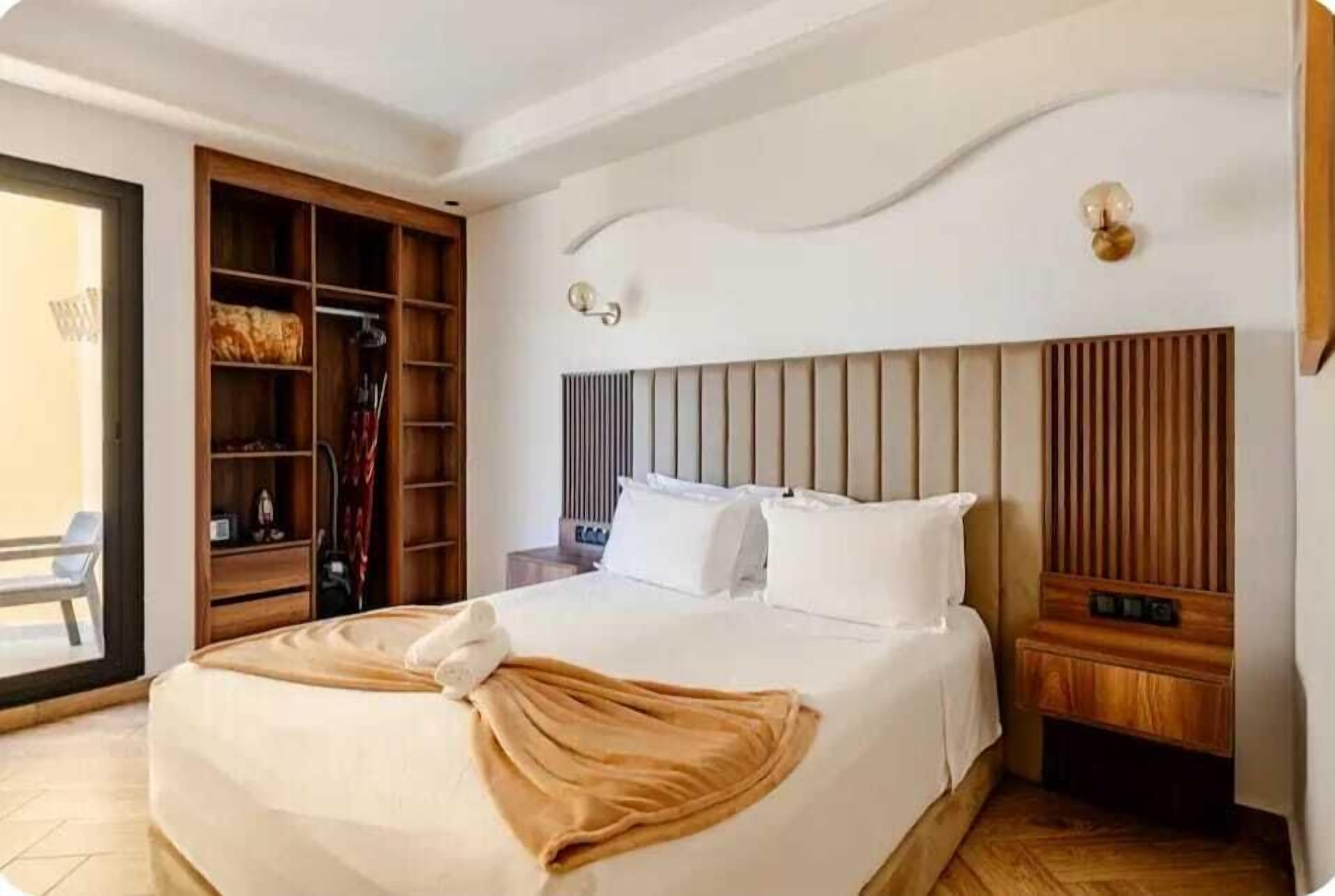 A hotel room with a bed, a wooden wardrobe, a bedside table, and a wall-mounted light fixture.