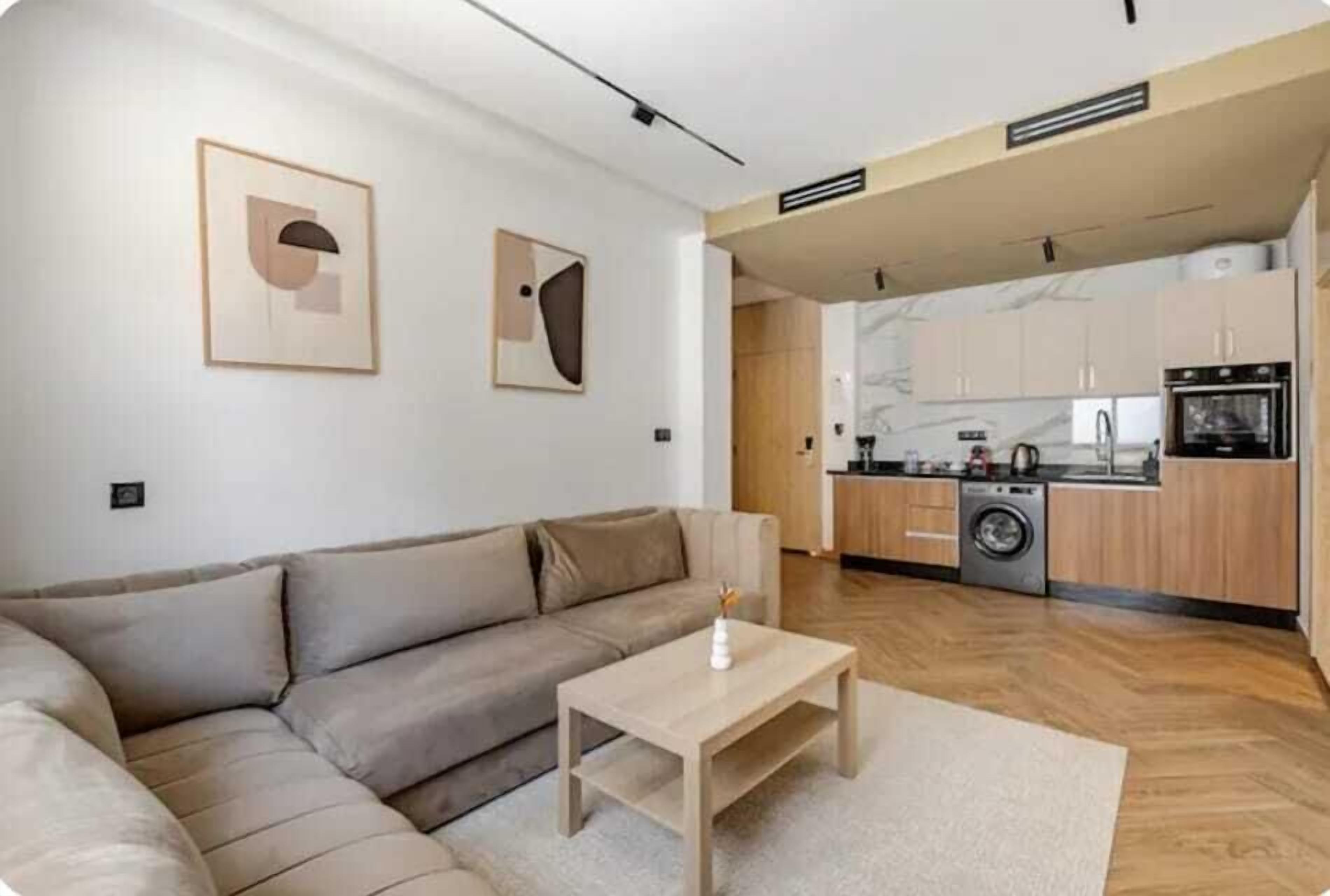 A modern living room with a sofa and a coffee table, a kitchen with wooden cabinets and appliances, and artwork on the walls.