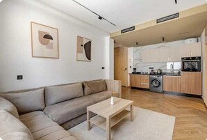 Una sala de estar moderna con un sofá y una mesita de centro, una cocina con armarios de madera y electrodomésticos, y obras de arte en las paredes.