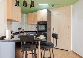Cocina compacta con protector mural de mármol, armarios de madera, encimera negra y taburetes de bar.