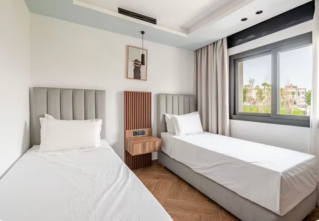 Una camera d'albergo con due letti, un comodino in legno, un quadro incorniciato e una finestra con tende.