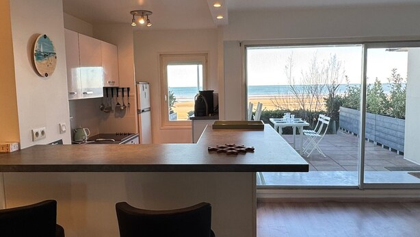 A modern kitchen with a large island, a dining area with a view of the beach, and a sliding glass door leading to an outdoor patio.