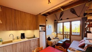 Une cuisine compacte avec des meubles en bois, un four à micro-ondes et un réfrigérateur. On y trouve un coin repas avec une table et des chaises rouges, ainsi qu’une fenêtre donnant sur des arbres et des montagnes.