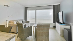 Ein modernes Hotelzimmer mit einer Sitzecke, zwei Korbstühlen, einem Esstisch, einem Flachbildfernseher und einem großen Fenster mit Vorhängen.