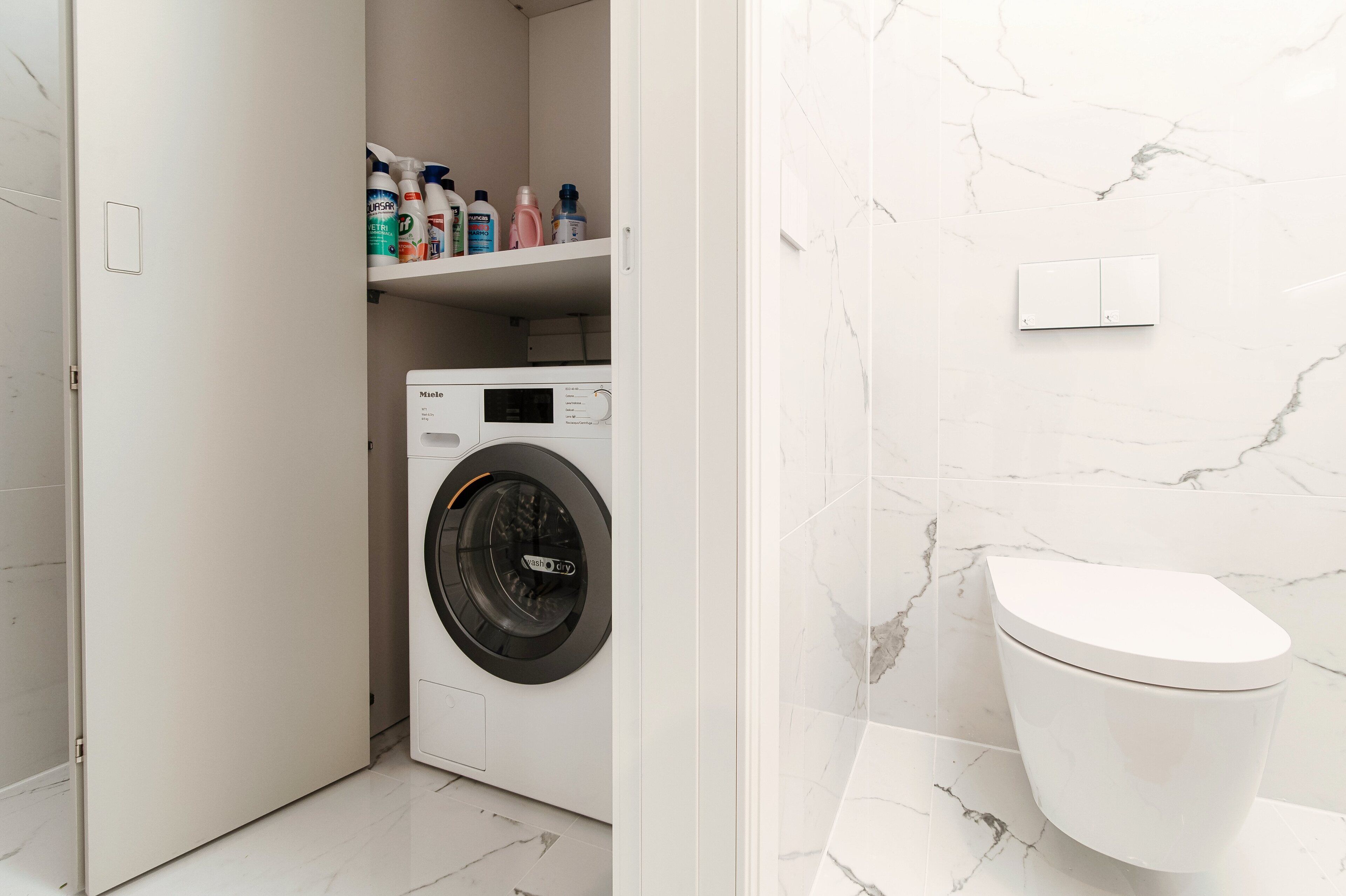 Une salle de bain moderne et compacte, dotée d’un lave-linge et d’un sèche-linge intégrés, de toilettes et d’une étagère contenant divers produits de toilette.