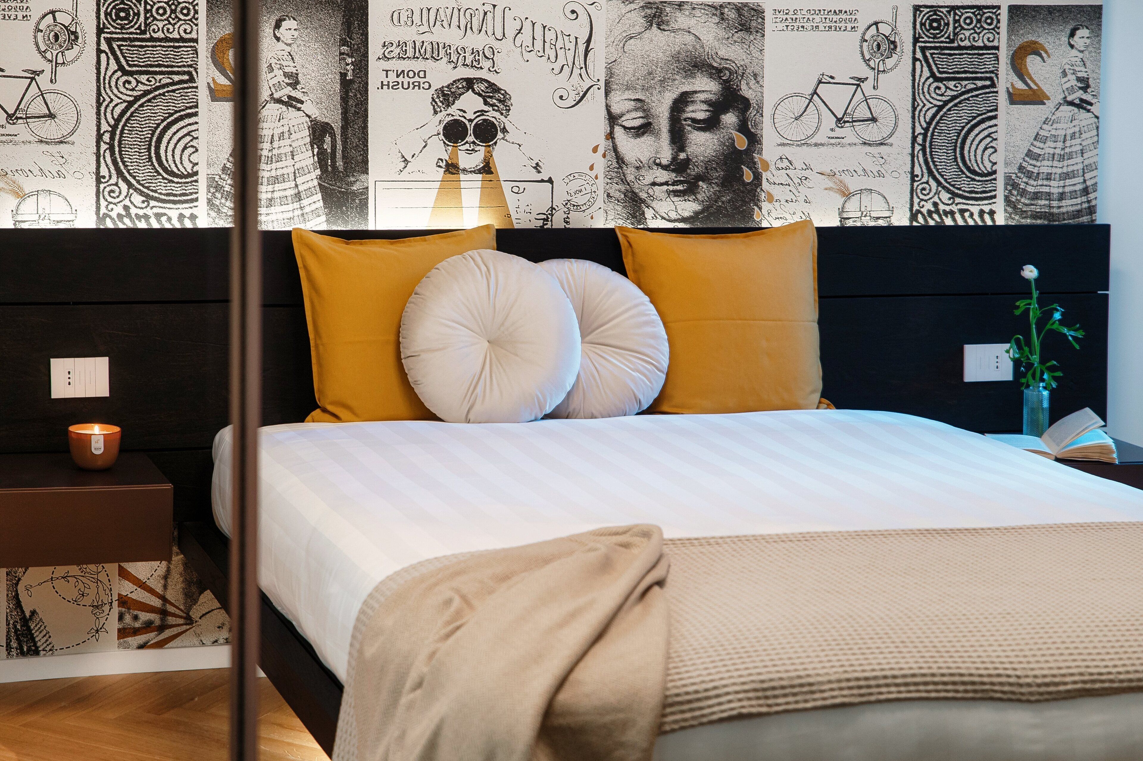 Una cama bien hecha con ropa de cama blanca y beige, dos almohadas grandes y una manta beige. Se observa una mesita de noche pequeña con una vela y un libro. La pared detrás de la cama presenta un papel tapiz blanco y negro con ilustraciones y texto.