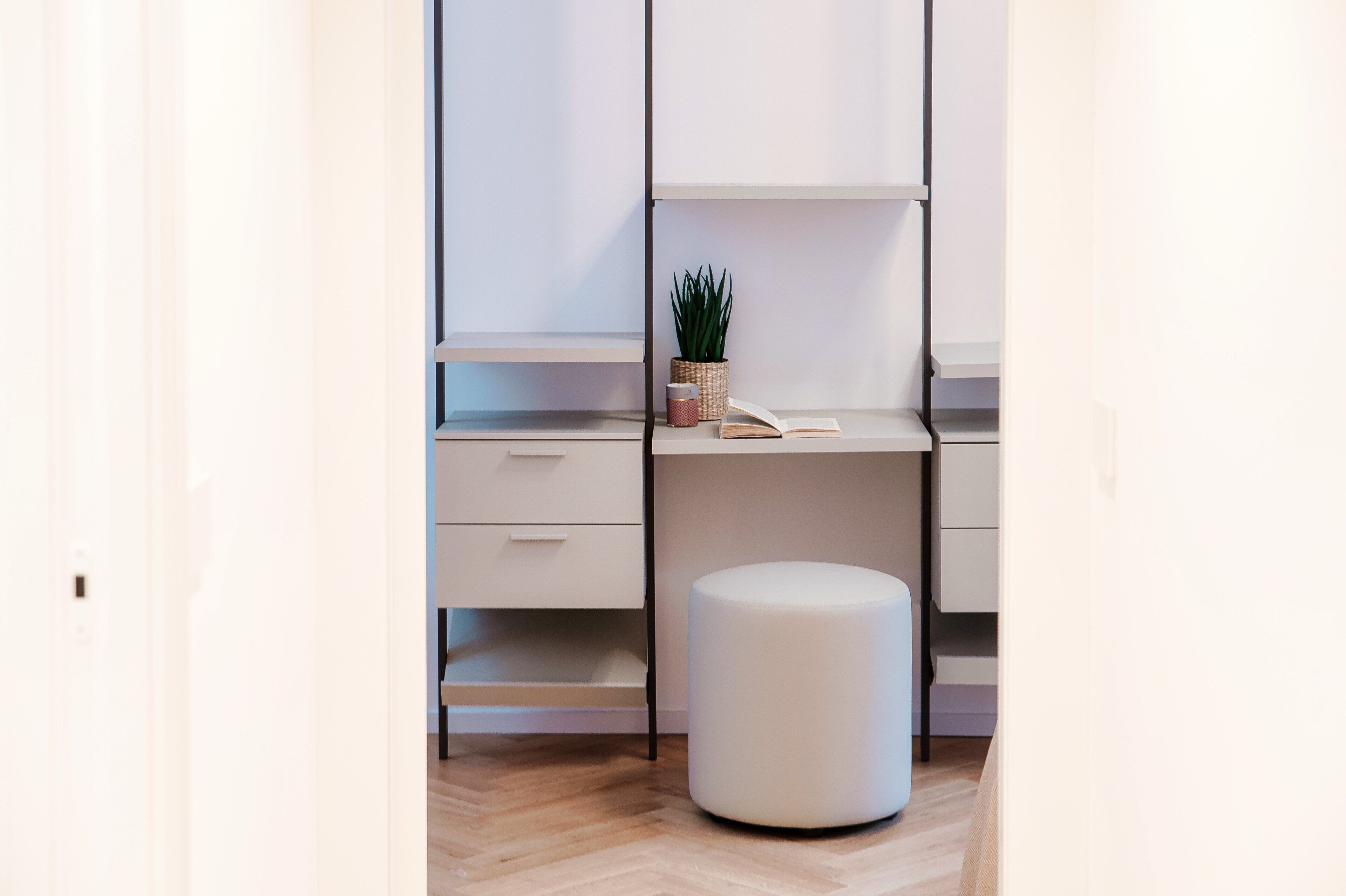 A modern, minimalist room with a wooden floor, a white desk with a plant, a wicker basket, and a small stool.