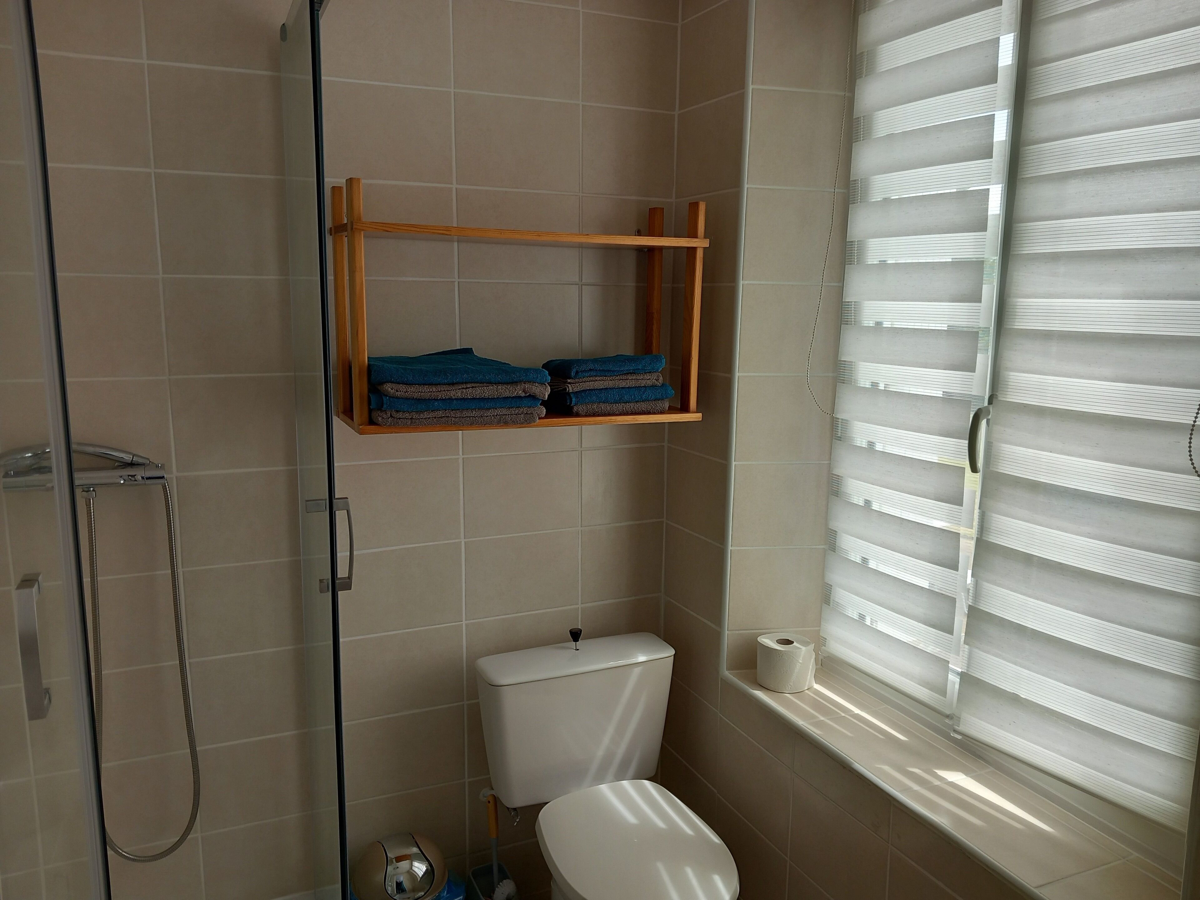 Une salle de bain comprenant des toilettes, une douche, une étagère avec des serviettes et une fenêtre avec des stores.