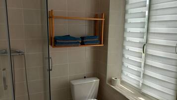 Une salle de bain comprenant des toilettes, une douche, une étagère avec des serviettes et une fenêtre avec des stores.