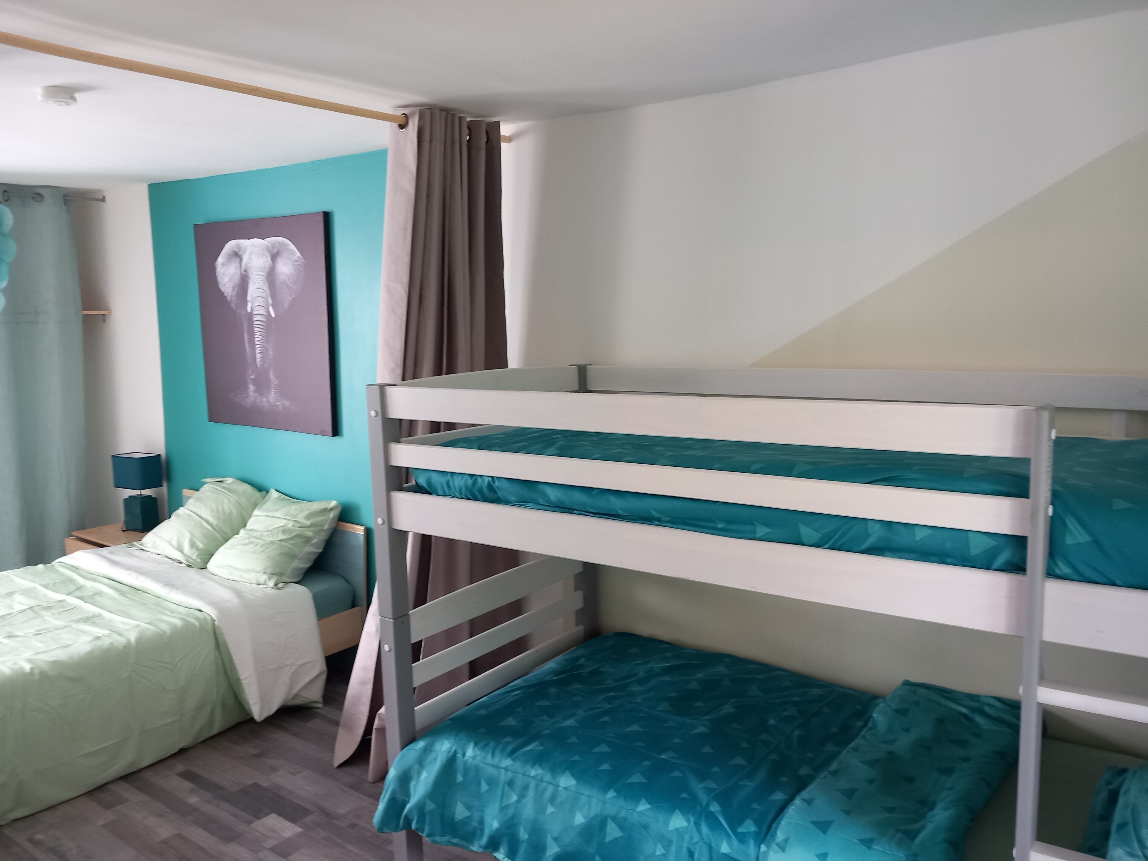 Un lit superposé avec une couette turquoise, une table de chevet avec une lampe, un lit avec une couette verte, un mur turquoise et une affiche d’un éléphant.