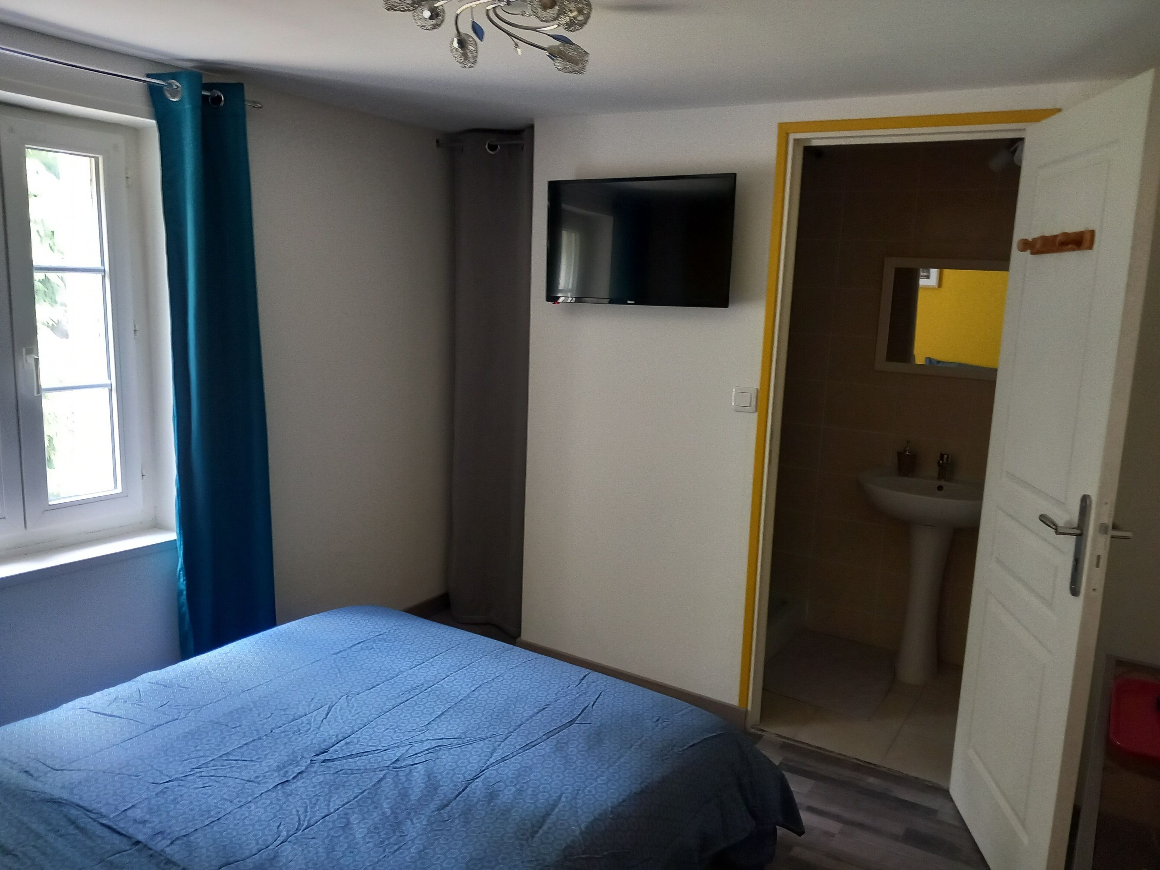 A bedroom with a bed, a TV mounted on the wall, a bathroom with a sink, and a door leading to another room.