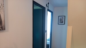 A hallway with a blue door leading to a room with a bed and a trash bin. - 3 BEDROOMS Villedieu les poeles MONT SAINT MICHEL (Sainte-Cécile)