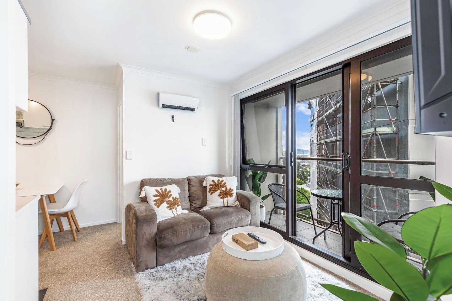 Ein modernes Wohnzimmer mit einer Couch, einem runden Hocker, einem Essbereich mit Tisch und Stühlen und einer großen Glasschiebetür, die auf einen Balkon führt.
