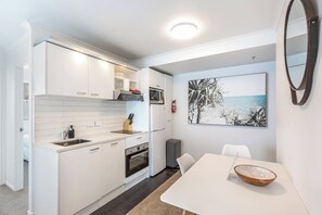 A compact kitchen with white cabinetry, a built-in oven, and a microwave. There's a dining area with a table and chairs, and a painting of a beach scene on the wall. - Central 3BR w/ Full Kitchen & Amenities by Zodiak (Auckland)