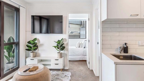 A compact, modern apartment with a kitchenette, a small seating area with a white sofa, and a large window with blinds. - Central 3BR Full Kitchen & Amenities Zodiak Stays (Auckland)