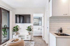 A compact, modern apartment with a kitchenette, a small seating area with a white sofa, and a large window with blinds. - Central 3BR Full Kitchen & Amenities Zodiak Stays (Auckland)