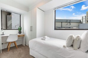 A modern bedroom with a large bed, a desk, and a window with a view of the sky and buildings. - Central 3BR w/ Full Kitchen & Amenities by Zodiak (Auckland)