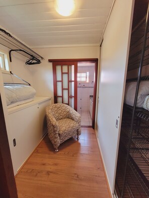 Un couloir étroit avec un plancher en bois, un fauteuil à motifs, un mur blanc orné d’un porte-serviettes et une porte donnant sur une autre pièce.