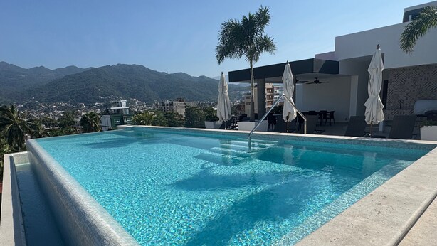 Ein rechteckiger Swimmingpool mit klarem, blauem Wasser, umgeben von einer Betonplattform. Weiße Sonnenschirme und Liegestühle sind vorhanden, und im Hintergrund ist eine Palme zu sehen.