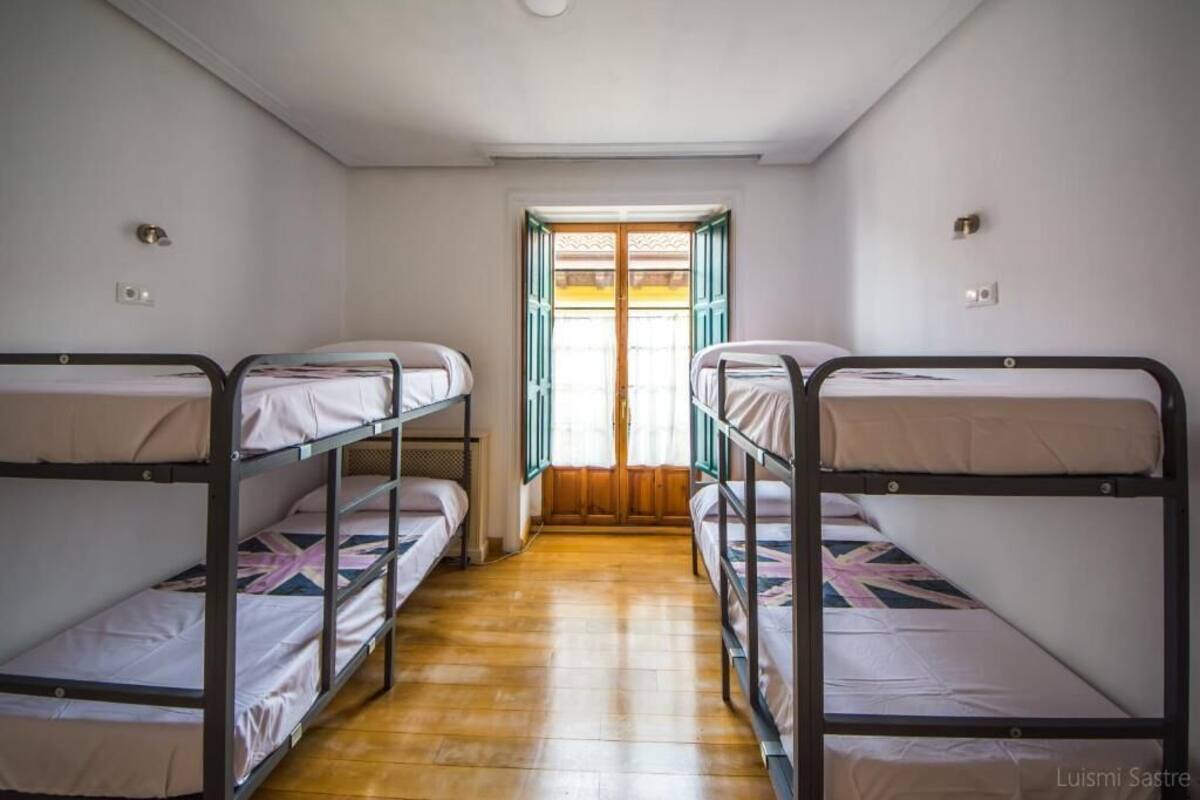 Una habitación con literas dispuestas en forma de U, suelo de madera y una ventana con cortinas.