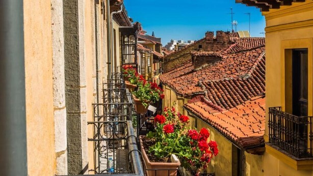 A narrow street with terracotta-tiled buildings, a balcony with red flowers, and a clear blue sky. - Hostel Covent Garden by gaiarooms  (León)