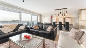 Ett modernt vardagsrum med ett soffbord i glas, svarta lädersoffor och en vit soffa. Det finns en matplats med ett bord och stolar, och ett kök syns i bakgrunden.