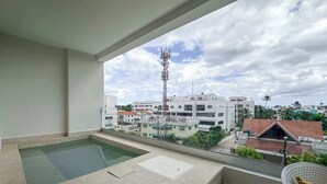 Un balcón con piscina, una silla y vistas a una zona residencial con edificios y una alta torre de comunicaciones.